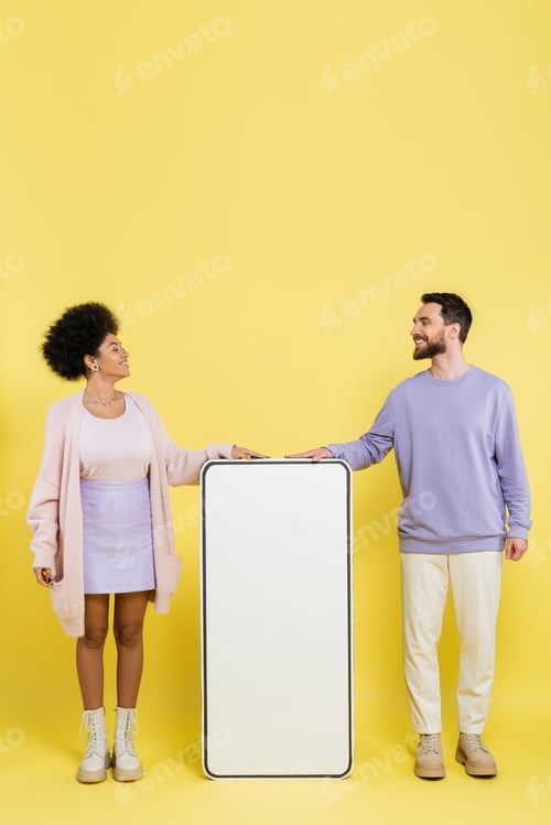 Preview: full length on trendy interracial couple smiling at each other near white mock-up of mobile phone on