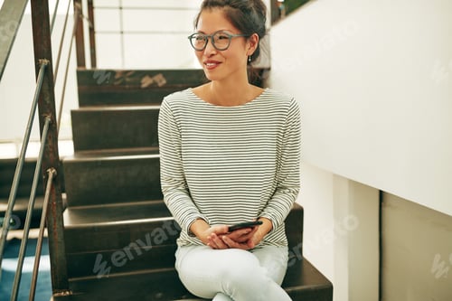 Preview: Asian businesswoman smiling while sitting on office stairs reading a text