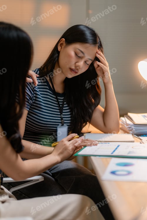 Preview: Stressed Asian businesswoman having headache at office desk, receiving support from colleague