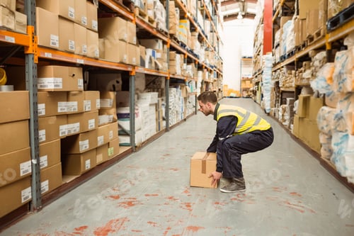 Preview: Male worker wearing high-visibility vest crouching and lifting cardboard box in storage aisle
