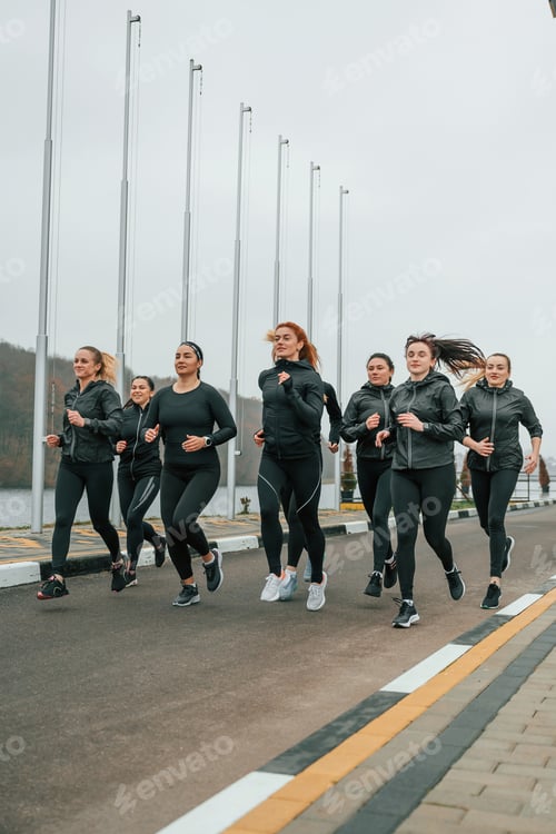 Preview: Running together. Group of sportive women is outdoors near black building