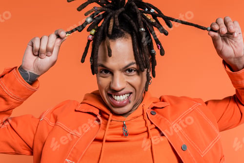 Preview: happy and tattooed multiracial man pulling dreadlocks isolated on coral