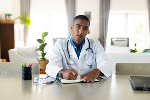 Preview: Biracial male doctor sitting at desk having video call, making notes in sunny room, slow motion