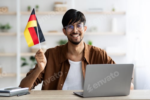 Preview: Smiling indian guy student showing flag of Germany, using laptop