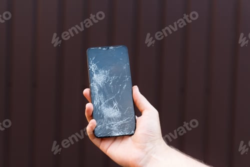 Preview: Broken glass screen smartphone in hand on brown background. Need for repair and the fragility of mob