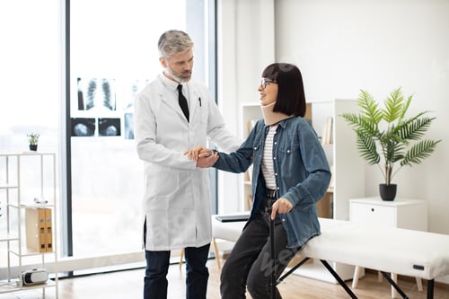 Preview: Specialist helping woman in leaning on cane in exam room