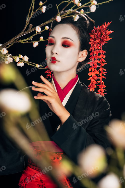 Preview: selective focus of beautiful geisha in black kimono with red flowers in hair and sakura branches