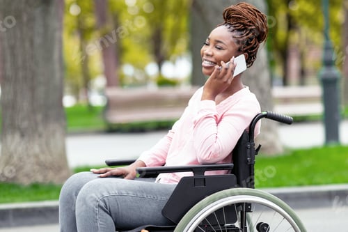 Preview: Beautiful young black lady in wheelchair speaking on smartphone at city park in autumn