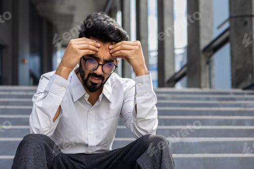 Preview: An upset and disappointed young Indian man sits on the steps of a business center and a courthouse