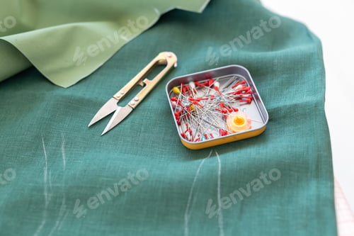Preview: Close up safety pins and sewing scissors lie on a green piece of linen fabric with markings. Concept