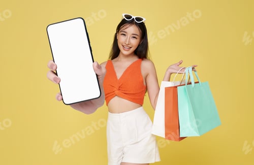 Preview: Happy young asian woman dressed in swimwear carrying shopping bag showing smartphone mockup
