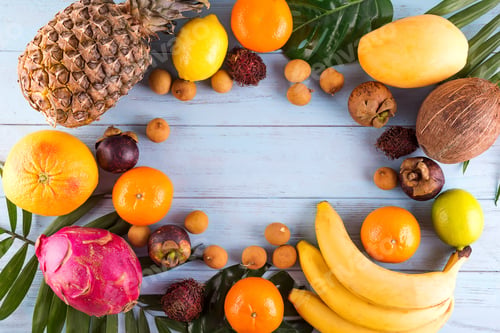 Preview: Healthy rainbow tropical fruits frame with palm leaves on blue wooden table, top view, copy space
