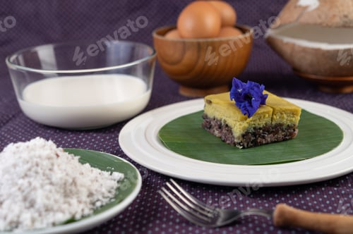 Preview: Black sticky rice and custard on a banana leaf in a white plate with butterfly pea flowers.
