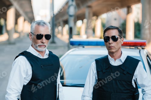 Preview: portrait of serious police officers in sunglasses and bulletproof vests near car at street