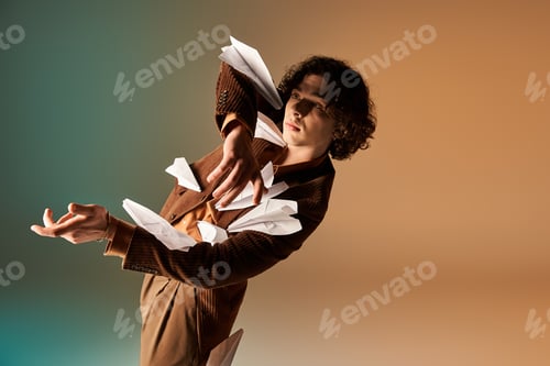 Preview: Curly haired young man showcases stylish flair with flying paper planes in studio
