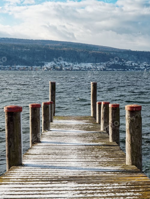 Preview: Wooden pier half covered in snow, with mountains and a small snowy village in the background.