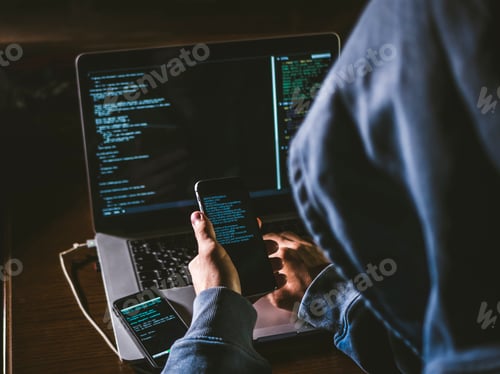 Preview: anonymous person in the hood sitting in front of computer working with laptop and a mobile phone