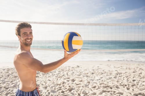 Preview: Athletic male standing on sandy beach holding volleyball near net under clear sky, copy space