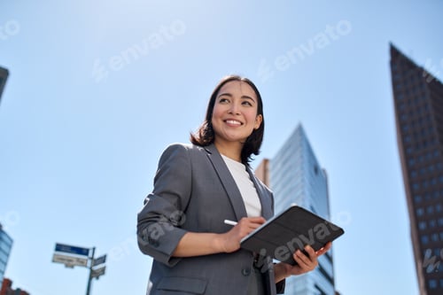 Preview: Smiling young Asian businesswoman using digital tablet on city street.