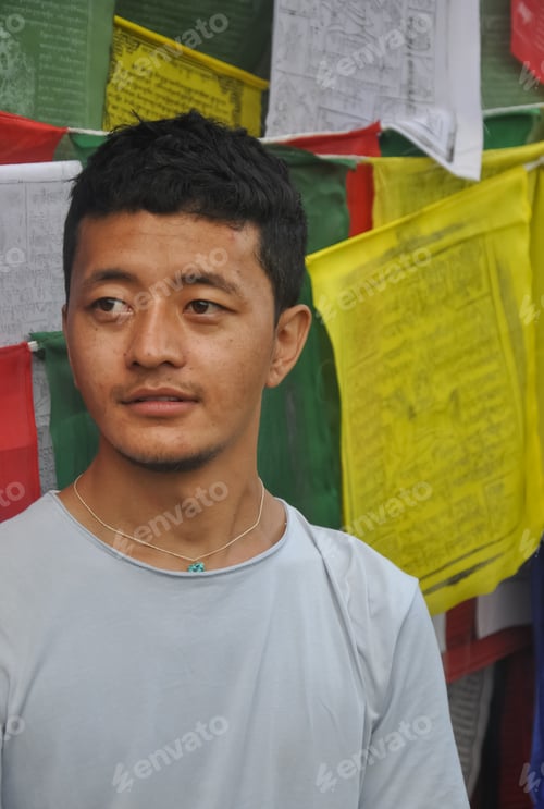 Preview: Closeup of a Ladakhi young guy looking sideways, standing against Buddhist prayer flags