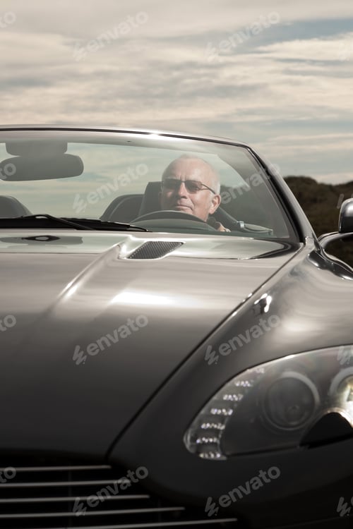 Preview: Senior Good Looking Happy Relaxed Retired Man With His Sports Car In Dune Landscape With Cloudy