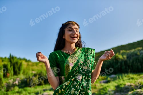 Preview: happy young indian woman in stylish sari posing and standing with landscape on background