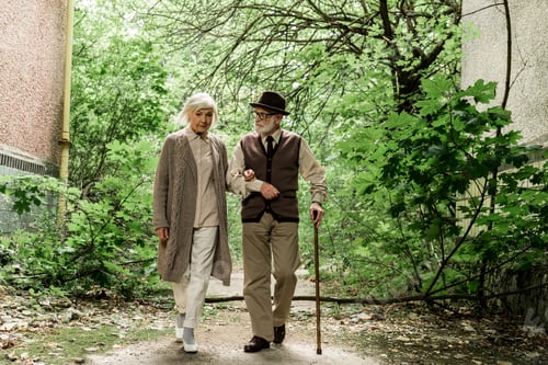 Preview: retired woman and senior man with walking cane walking near green trees
