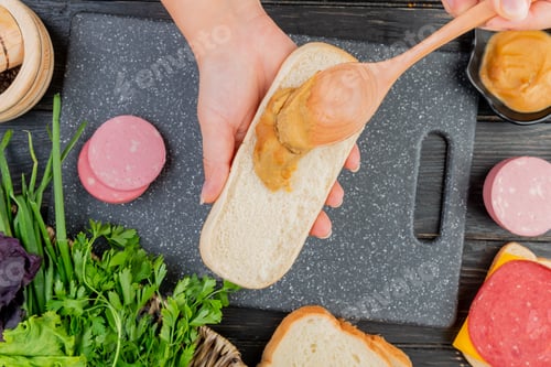 Preview: top view of female hands spreading mustard on sandwich with spoon and boiled sausage