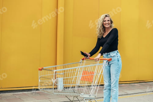 Preview: Woman using smartphone while holding shopping cart in front of the supermarket or the mall
