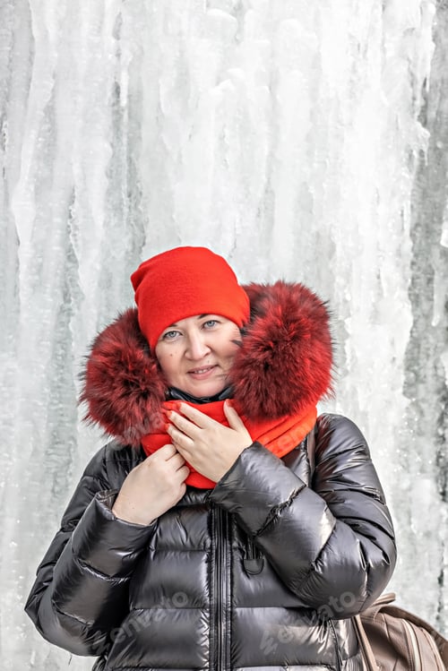 Preview: Portrait of a woman in a red hat and scarf, warm jacket against the background of an ice wall.Winter