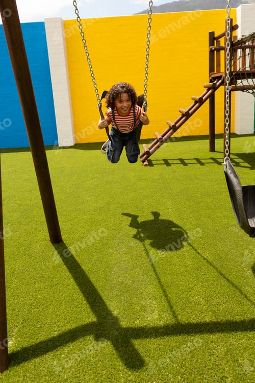 Preview: Front view of mixed-race schoolgirl playing on a swing at school playground on a sunny day