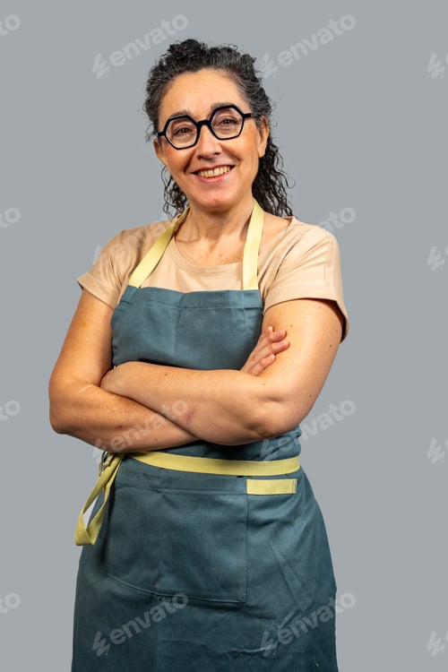 Preview: Smiling senior woman wearing apron and glasses with crossed arms on gray background
