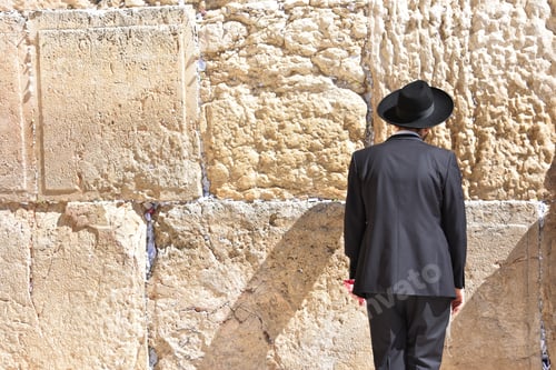 Preview: Man standing in a suit and a hat looking at a rock wall in the sun