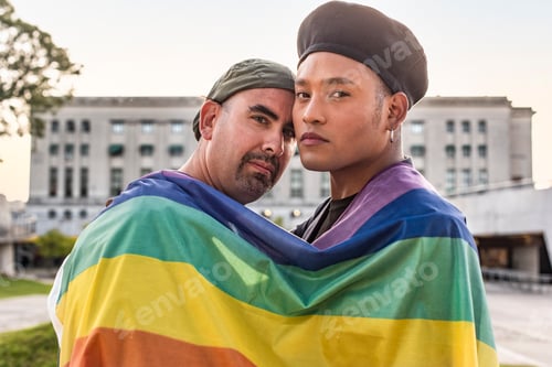 Preview: Gay couple under LGBT rainbow flag while looking at camera