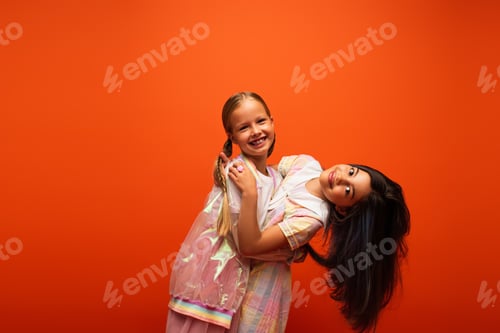 Preview: cheerful girl embracing brunette friend while smiling at camera isolated on orange