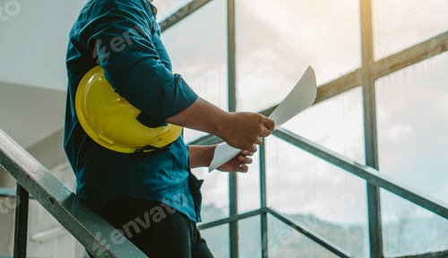 Preview: Architect man holding hardhat and blueprint while standing outdoors and against building structure.