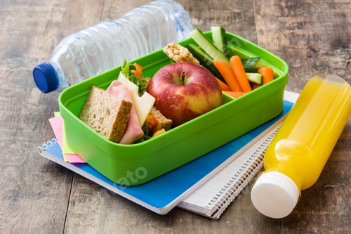 Preview: Lunchbox with sandwich, apple, vegetables and juice resting on notebooks
