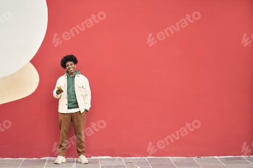 Preview: Happy young African American guy standing at color wall outdoors using mobile.
