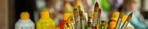 Preview: Close up of artists paintbrushes and bottles of paint against a blurred crowd.