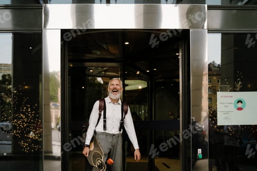 Preview: Grey mature man smiling while standing with skateboard by building