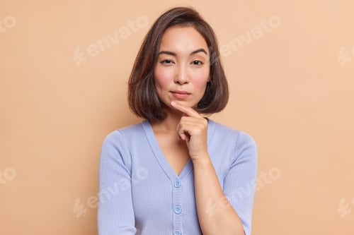 Preview: Portrait of serious dark haired Asian woman keeps finger on chin looks mysteriously at camera consid