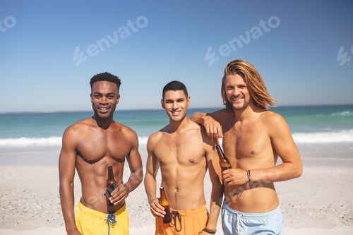 Preview: Portrait of young male diverse friends holding beer bottles and looking at camera on the beach
