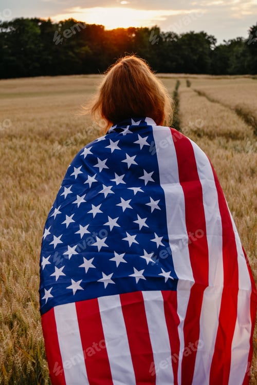 Preview: Woman walking with flag of USA in wheat field at sunset