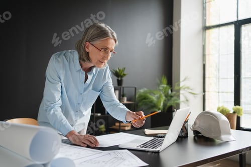 Preview: Mature Caucasian lady office desk with a laptop holding pencil working with blueprint floorplans