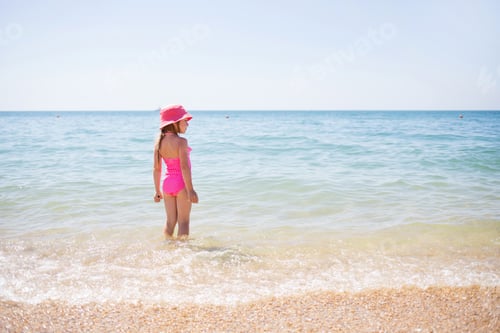 Preview: child girl in pink swimsuit and panama stands in water of sea on sunny summer day against background