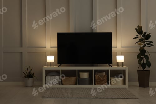 Preview: Modern TV on cabinet, lamps and beautiful houseplants near white wall in room. Interior design