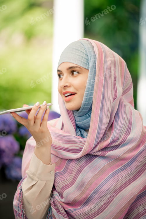 Preview: Young female in hijab recording voice message on smartphone outdoors