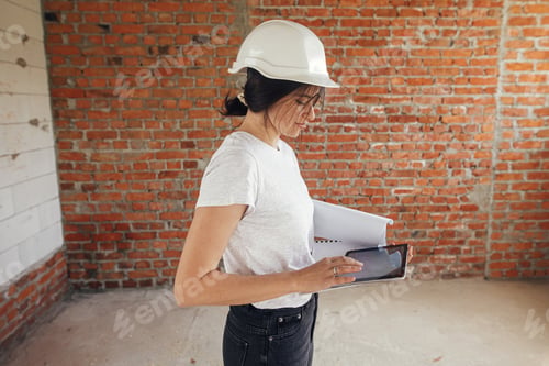 Preview: Stylish woman architect in hard hat with tablet checking blueprints at construction site