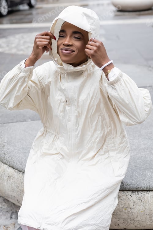 Preview: Young african american woman in raincoat sitting on blurred urban street