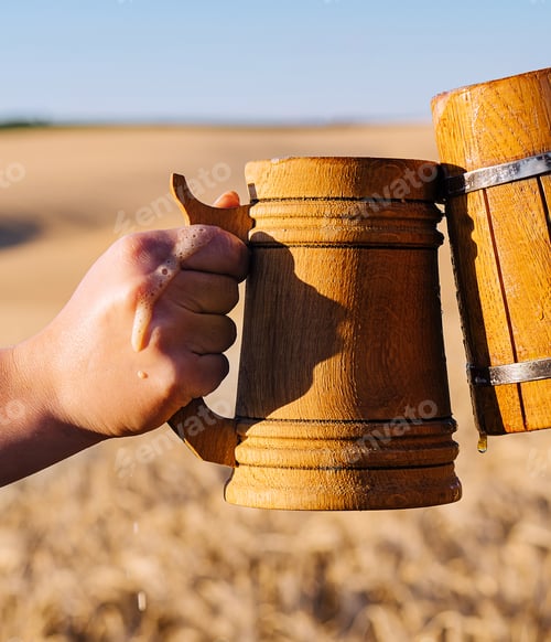 Preview: Hands with wooden beer clink mugs on the background of a field of wheat
